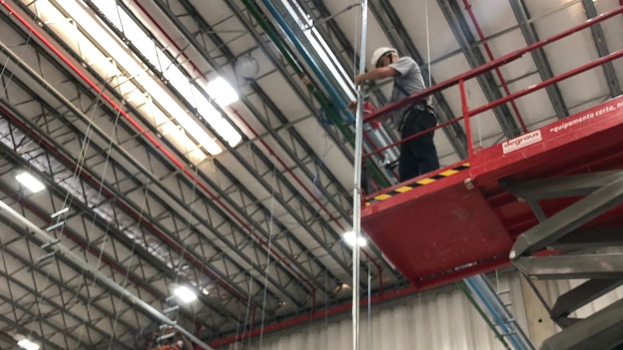 Técnicos instalando cabeamento em altura com plataforma elevatória (PTA) em galpão logístico, seguindo NR-35
