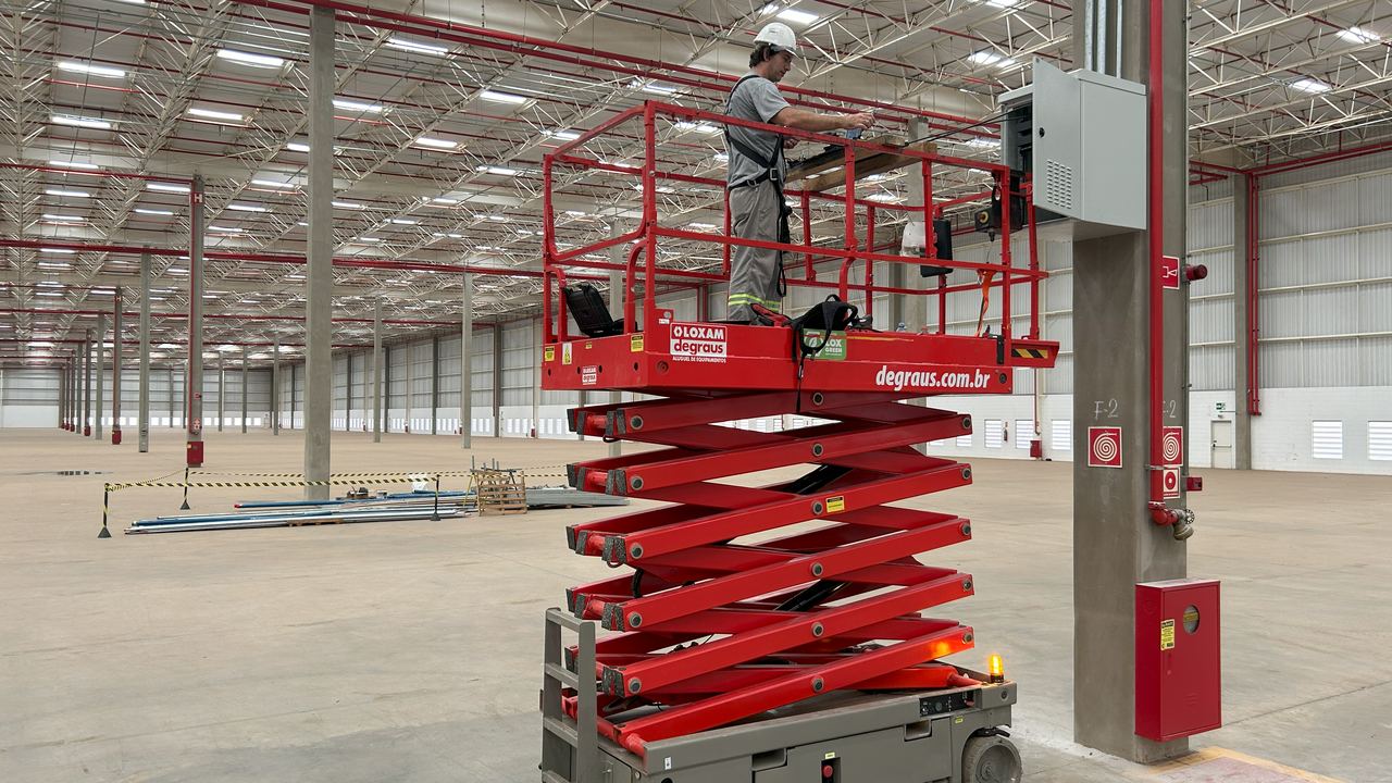 Técnico em plataforma elevatória tesoura, com cinto de segurança e capacete, instalando infraestrutura de rede em pé-direito alto
